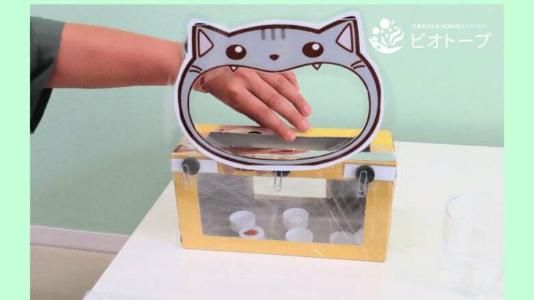 大きく口を開けたねこのイラストが貼ってある箱に、イチゴの絵を貼り付けたペットボトルキャップを使うおもちゃ。ネコの口に入れてあげる。